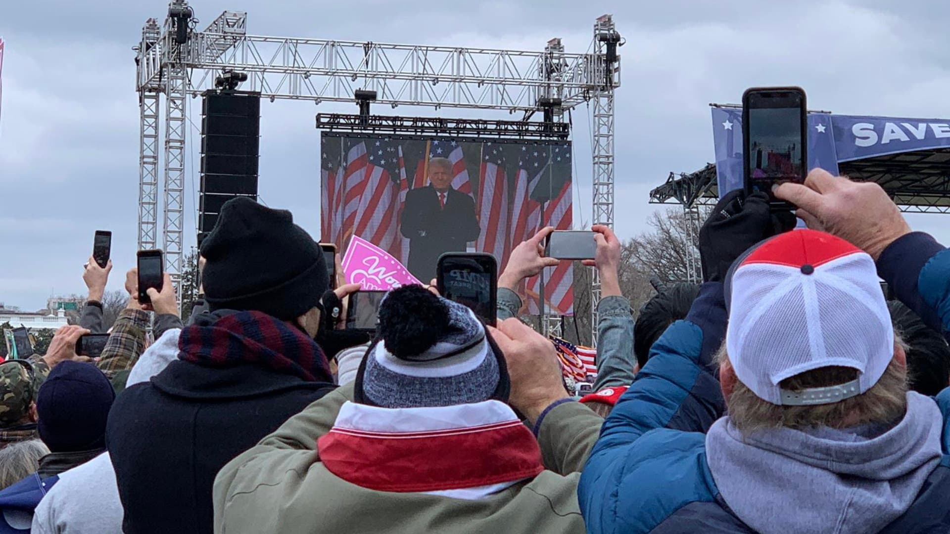 Crowd members hold up phones while Donald Trump speaks on a large screen at the January 6, 2021 rally in Washington.