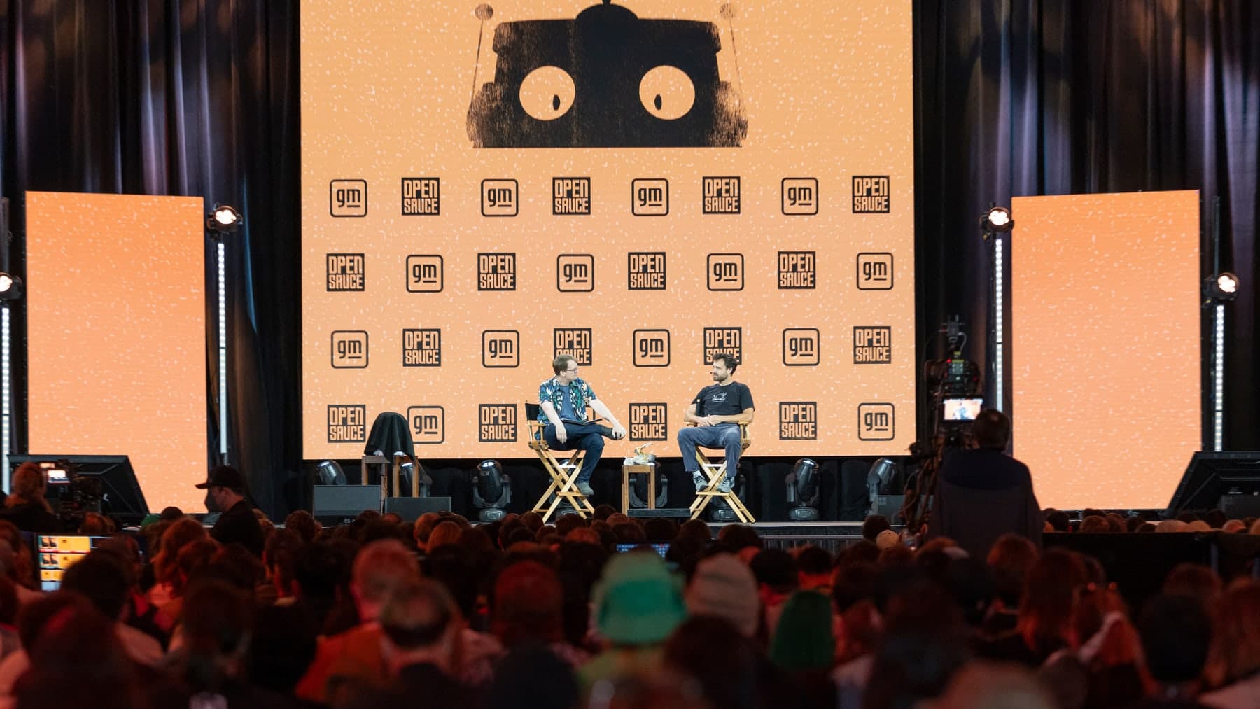 Hank Green and William Osman seated on a stage beneath a large illustrated robot head at Open Sauce 2026.
