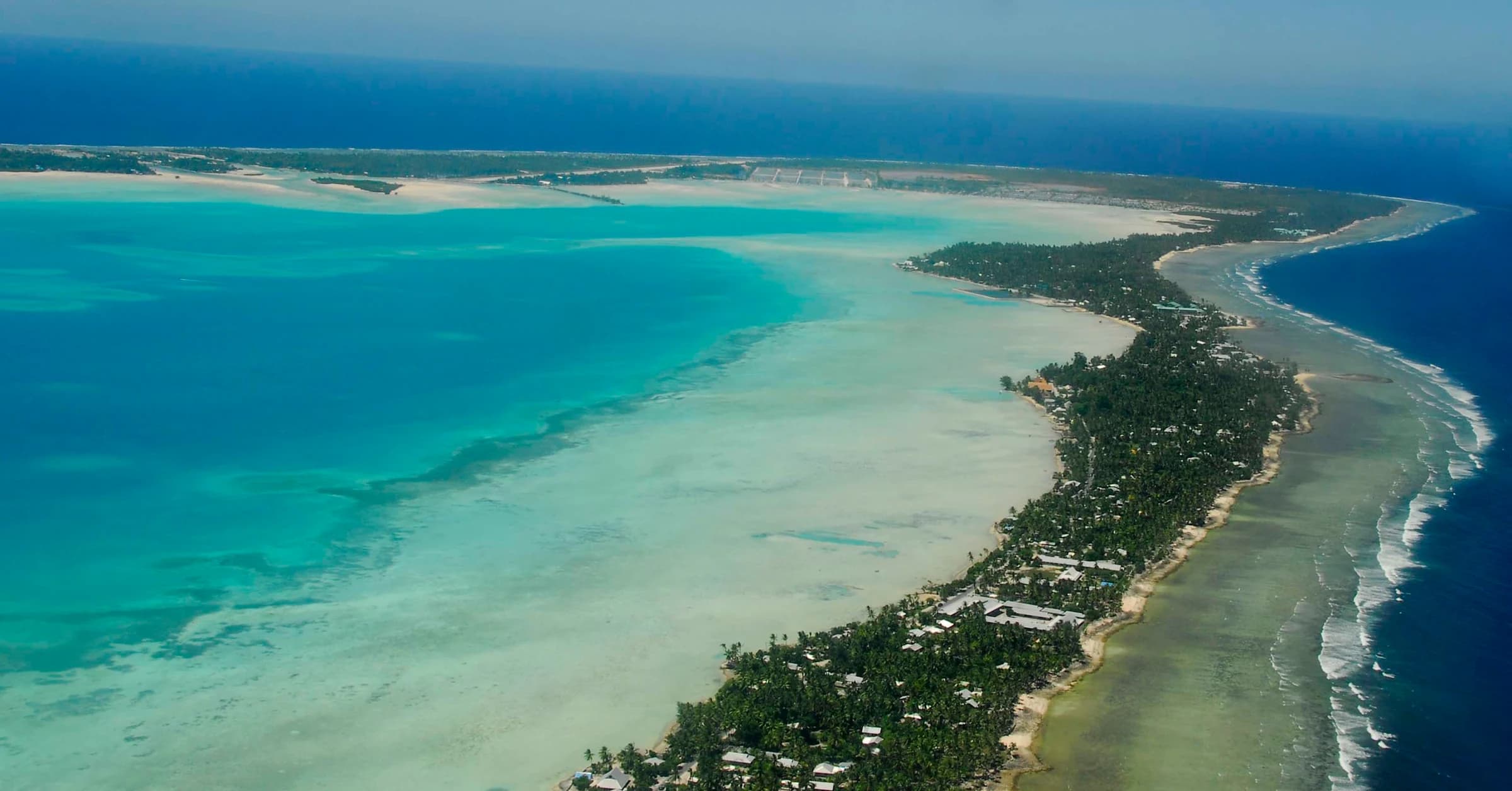 Aerial view of a narrow inhabited strip of South Tarawa between a turquoise lagoon and the Pacific Ocean.