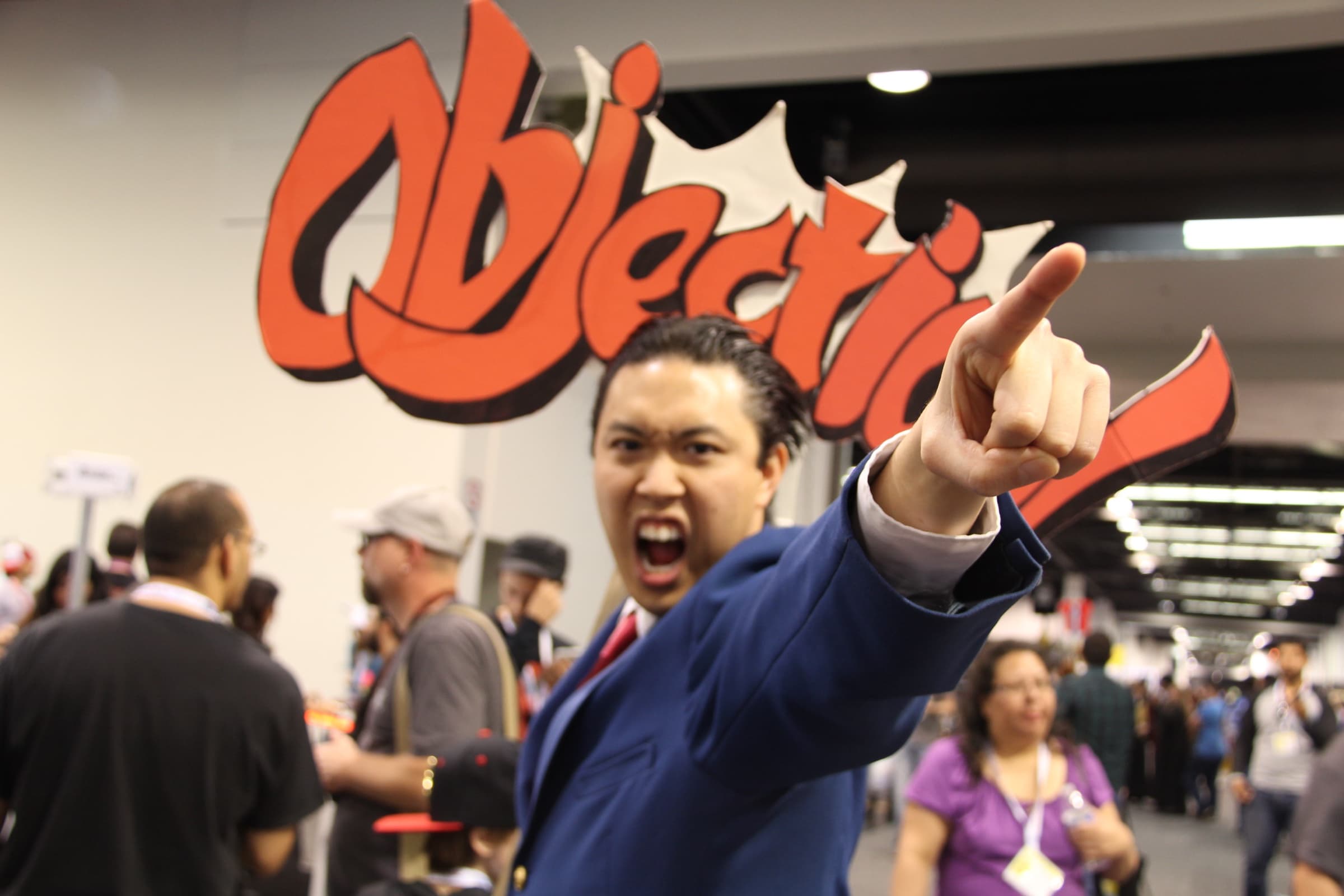 A Phoenix Wright cosplayer in a blue suit points beneath a large red Objection sign at WonderCon 2015.